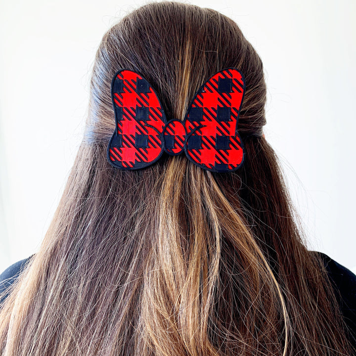 A person's head viewed from the side, with a buffalo plaid interchangeable bow hair clip in red and black checkered pattern, attached to a headband.